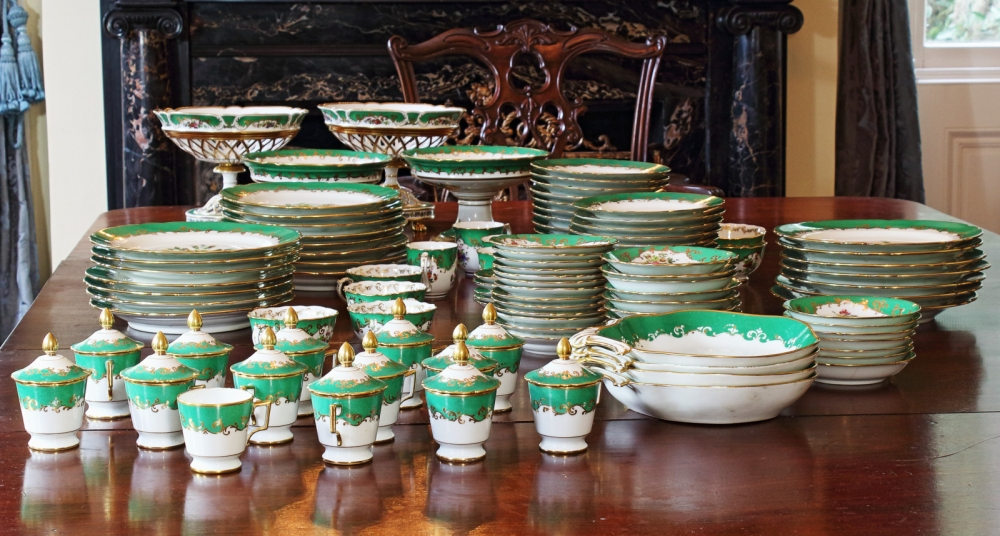 An ornate collection of china dishes is elegantly arranged on a polished wooden table. The set features green and gold detailing on white porcelain, including plates, bowls, and lidded containers. A carved chair is visible in the background.
