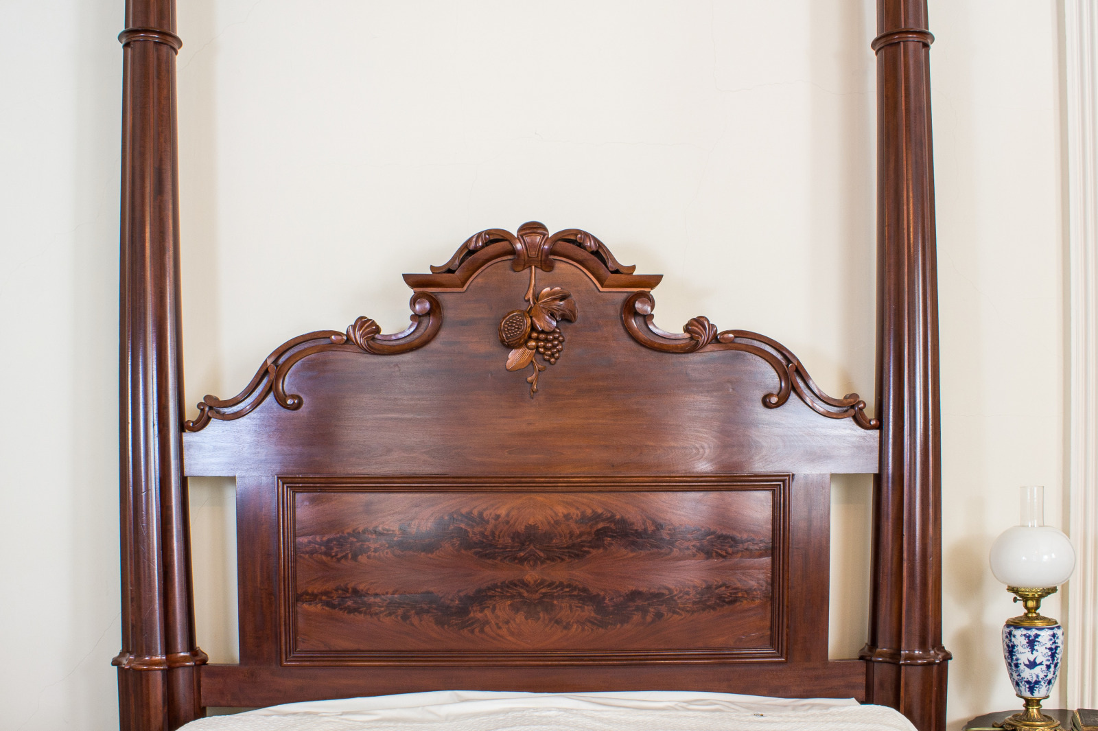 Antique wooden four-poster bed with an ornate, carved headboard featuring a fruit motif. A vintage oil lamp with a blue pattern sits on a nearby table against a cream wall.