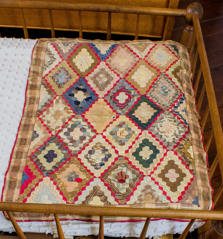 A colorful, vintage quilt with intricate patchwork patterns lies draped over a wooden crib. The quilt features diamond shapes in a variety of colors, bordered by a red trim.