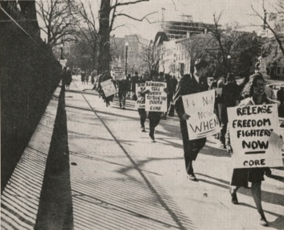 A black and white photo shows a group of protesters walking on a sidewalk, holding signs with messages such as Release Freedom Fighters Now and If Not Now, When? The scene is set near a fence with trees lining the path.
