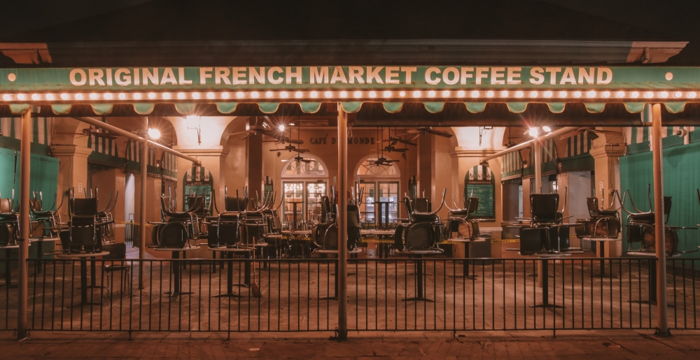 A dimly lit outdoor café with a green canopy reading Original French Market Coffee Stand. Chairs are stacked on tables behind a metal fence, indicating closed or after hours. Warm lights create a cozy atmosphere.