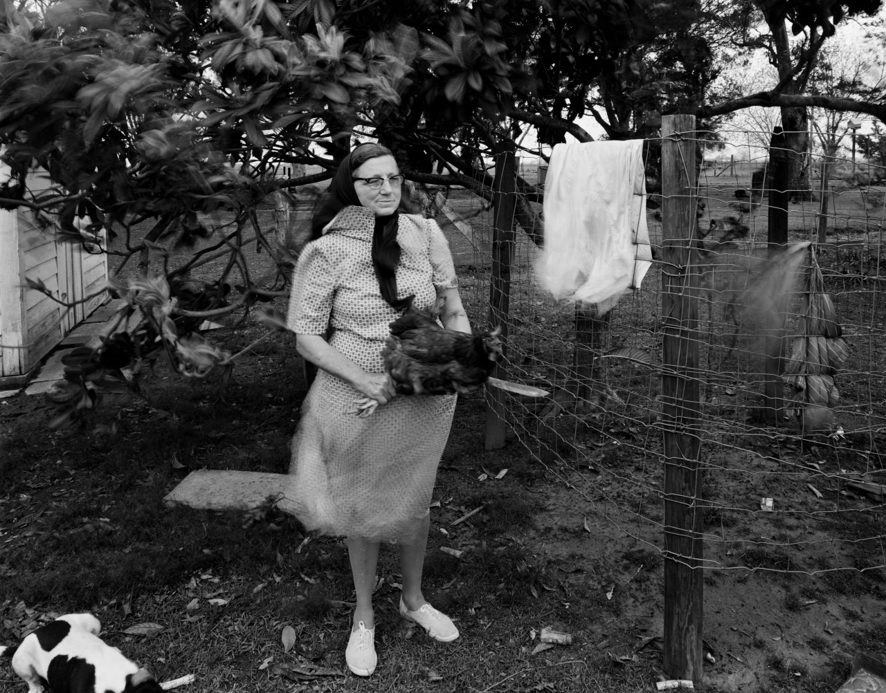 A black-and-white photo of a woman standing by a fence, holding a chicken. She is wearing a patterned dress and glasses, with trees and a cloth hanging on the fence in the background. A dog is lying on the grass in the foreground.