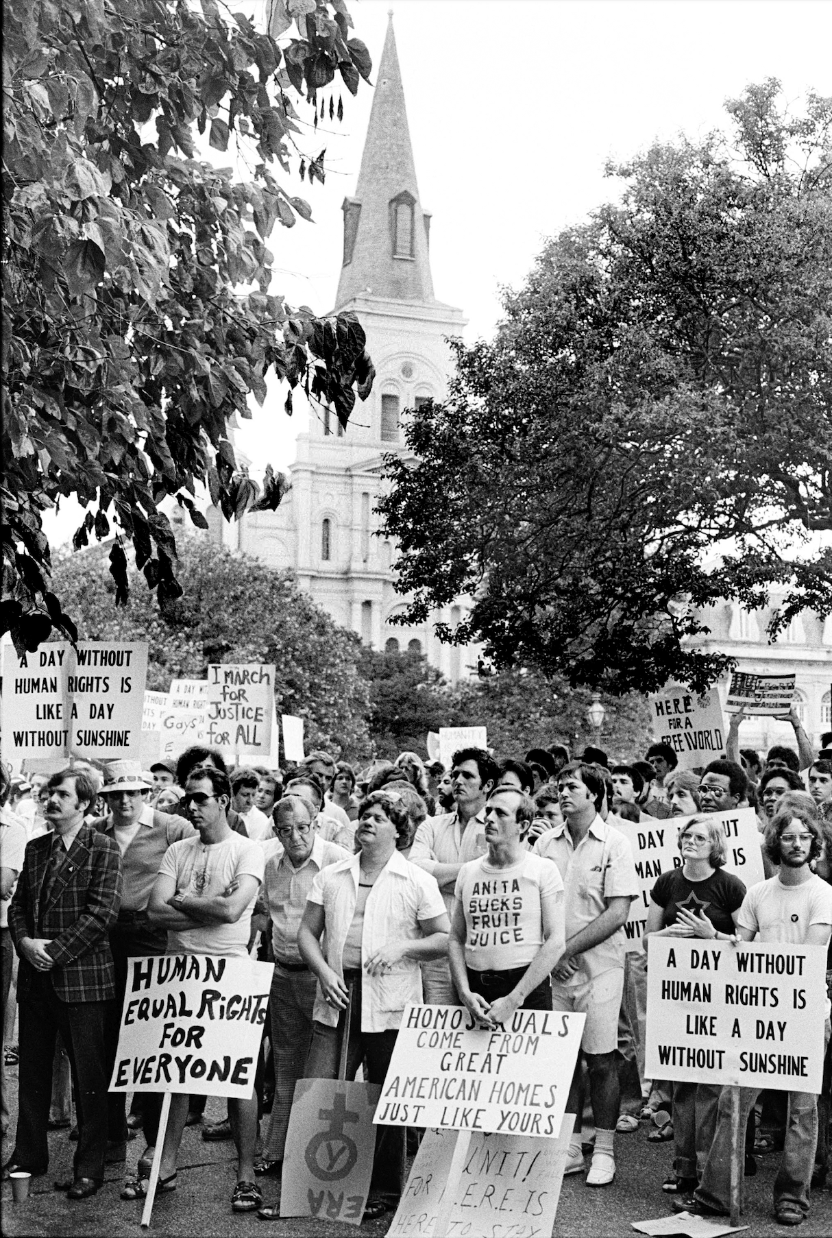 A black and white photo shows the HERE demonstration in Jackson Square on June 18, 1977. Protesters with signs are in the foreground with St. Louis Cathedral visible in the background.