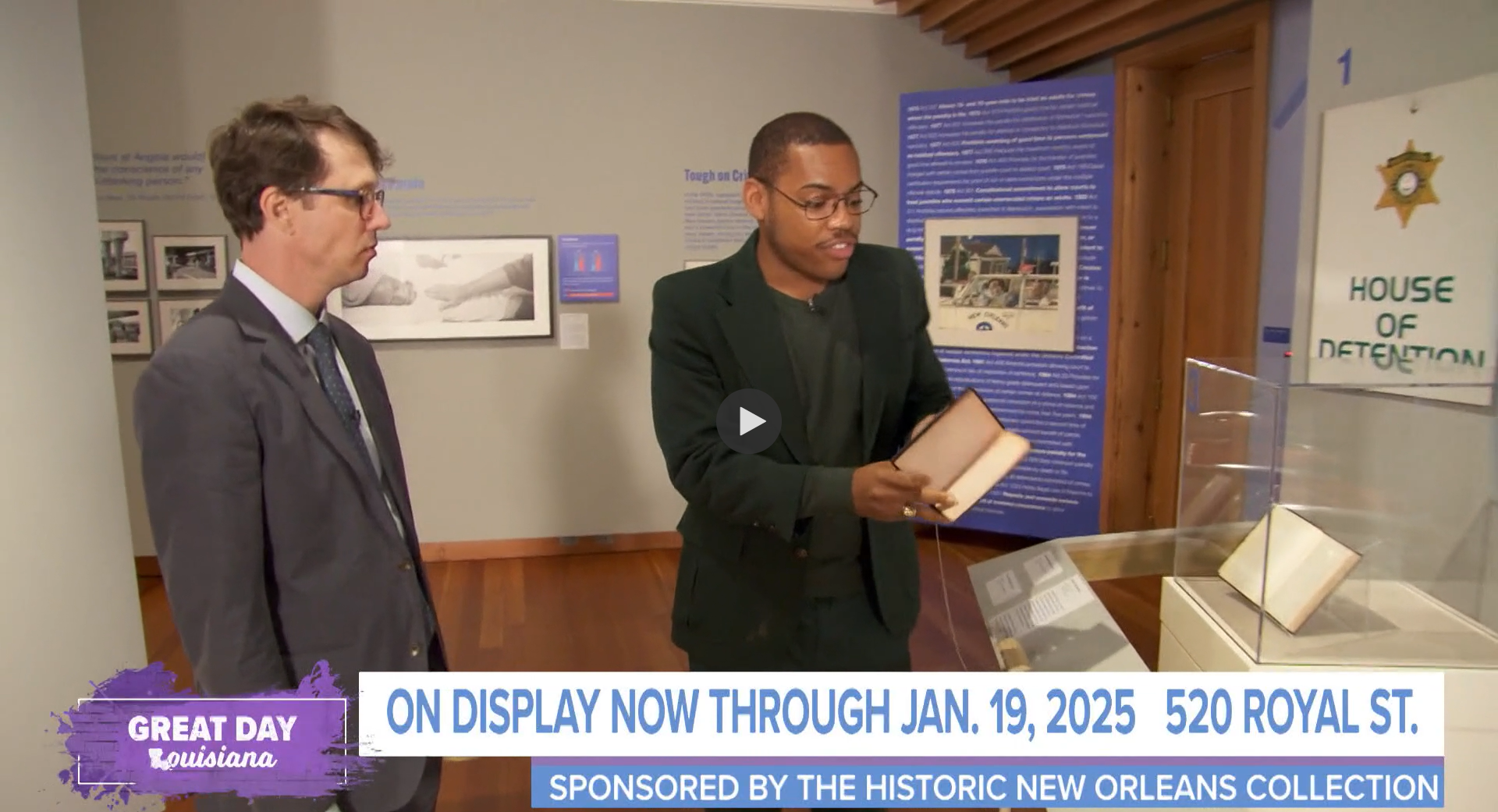 Two men are observing an exhibit in a museum. One holds a book while explaining something. Behind them are informational panels and photographs. Text overlays mention a display through January 19, 2025, at 520 Royal St., sponsored by the Historic New Orleans Collection.