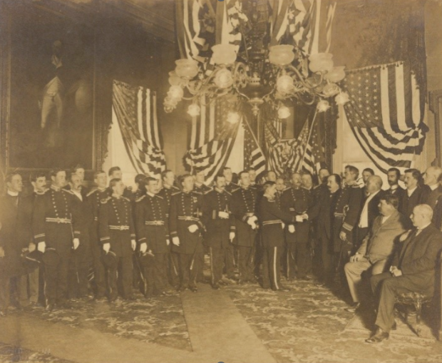 A vintage photo depicts a group of men in military uniforms standing in an ornately decorated room with large American flags draped in the background. Chandeliers hang above, and several men are seated on the right side of the image.