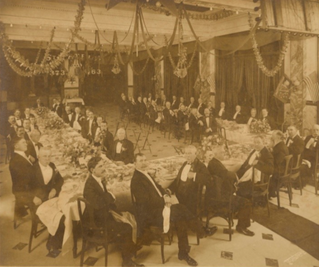 A vintage photograph depicting a formal banquet in an ornate hall. Numerous men in suits and bow ties are seated at long, lavishly decorated tables, surrounded by garlands hanging from the ceiling.