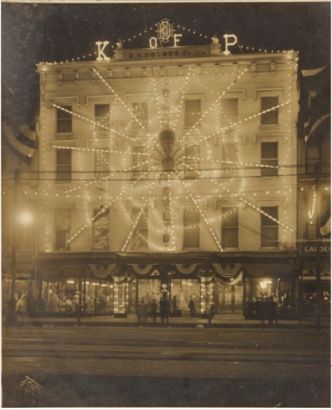 Sepia-toned image of a building decorated with elaborate lights forming a circular pattern. The facade features the letters K of P. Part of the street and people are visible in front of the building.