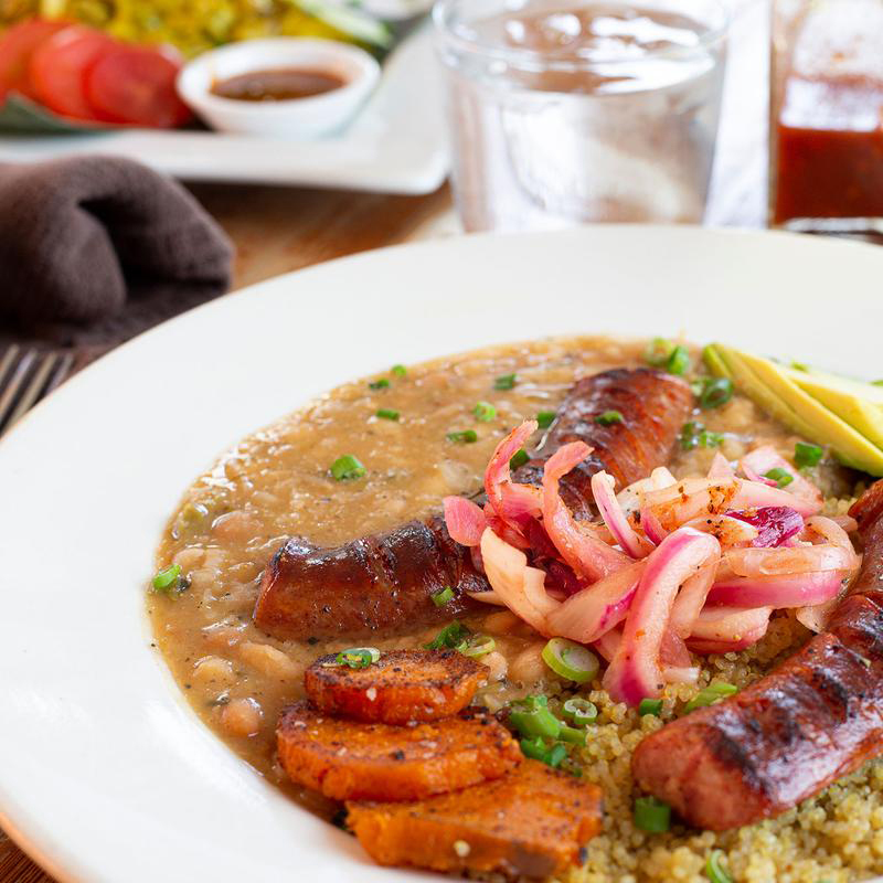 A plate of food featuring grilled sausages, quinoa, beans, and roasted sweet potatoes, garnished with pickled onions and avocado slices. A glass of water and a side dish are visible in the background.