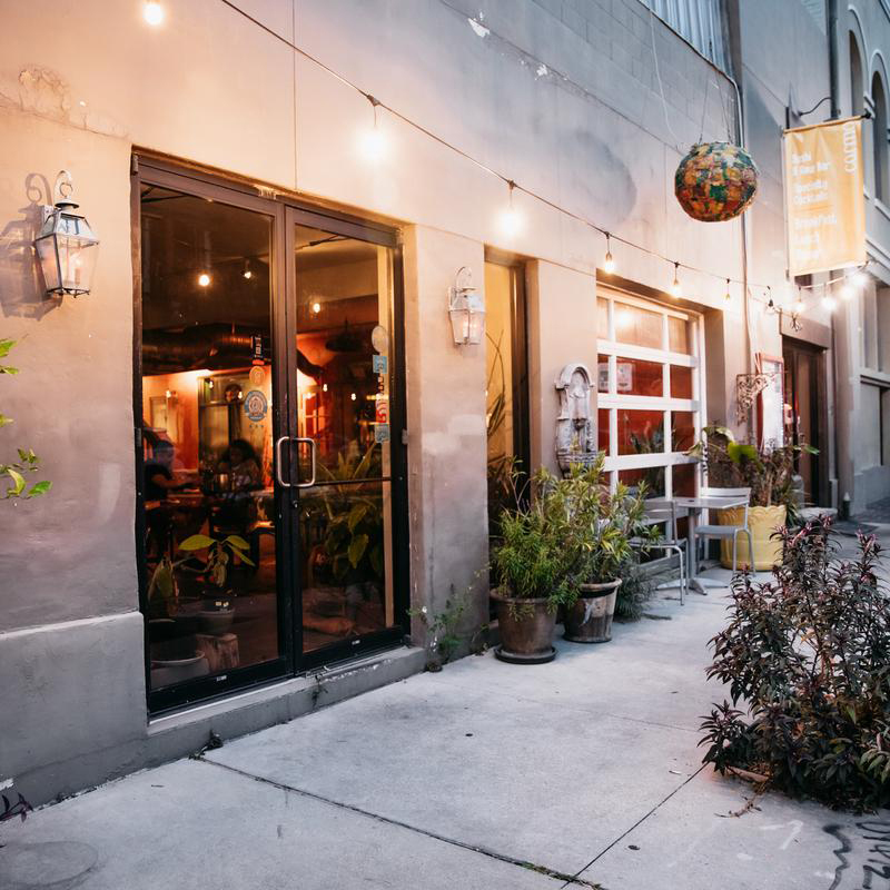 Exterior view of a cozy café with large glass doors and windows. The entrance is adorned with potted plants and lit by string lights. Tables and chairs are set up outside, and a warm glow emanates from inside.