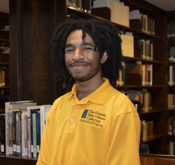 A person with long, curly hair is smiling while standing in a library. They are wearing a yellow polo shirt with The Historic New Orleans Collection Internship Program written on it. Shelves filled with books are in the background.