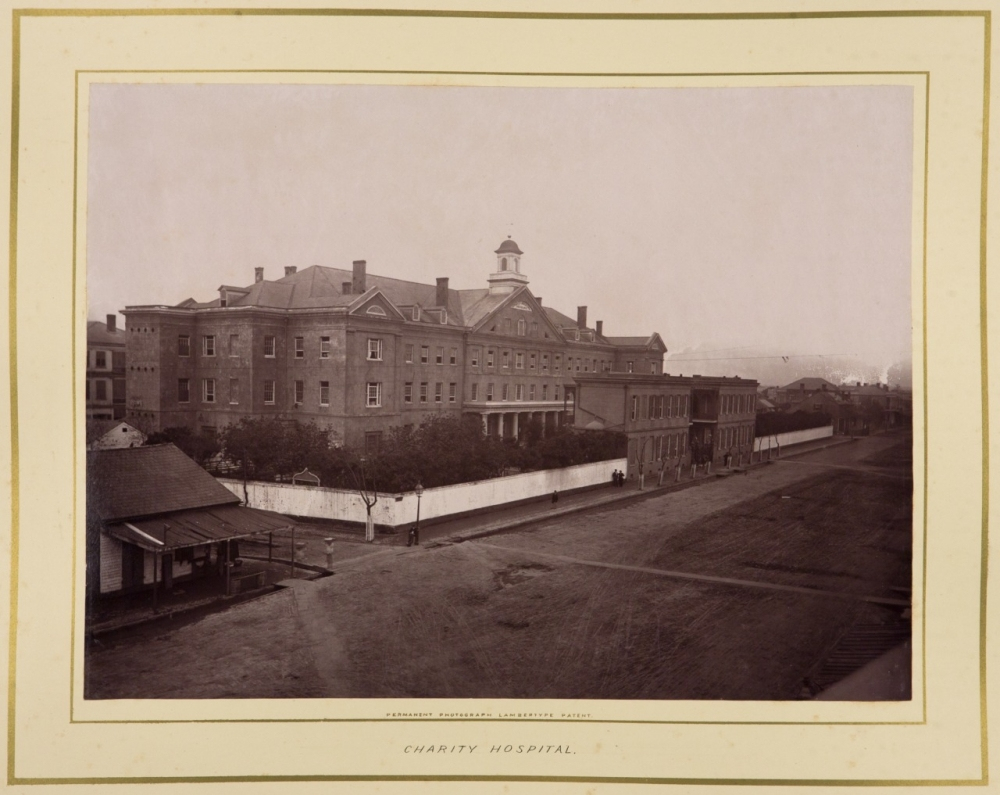 A historic black and white photograph of Charity Hospital, showcasing a large, multi-story brick building with a cupola. The foreground includes a dirt road and smaller adjacent structures. Trees line the property, enclosed by a fence.