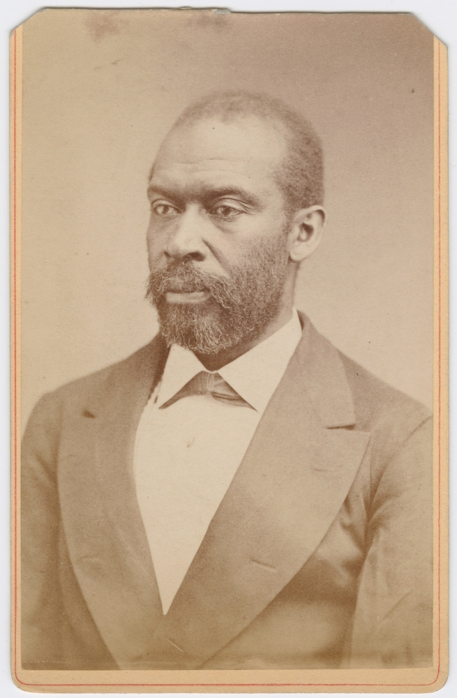 Sepia-toned portrait of a man in a suit with a bow tie, looking to the side. The image has a vintage appearance, with slight wear visible around the edges.