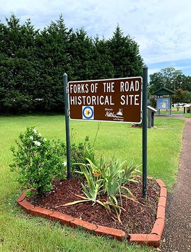 A brown sign reading Forks of the Road Historical Site with surrounding greenery, including bushes and small plants. The site is located next to a paved path with trees and a clear sky in the background.