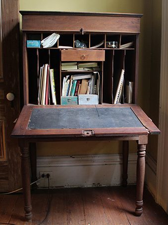 A vintage wooden secretary desk with a fold-out writing surface. It features open cubbies filled with books, papers, and small boxes, situated in a dimly lit room. The desk has turned legs and a warm, worn finish.