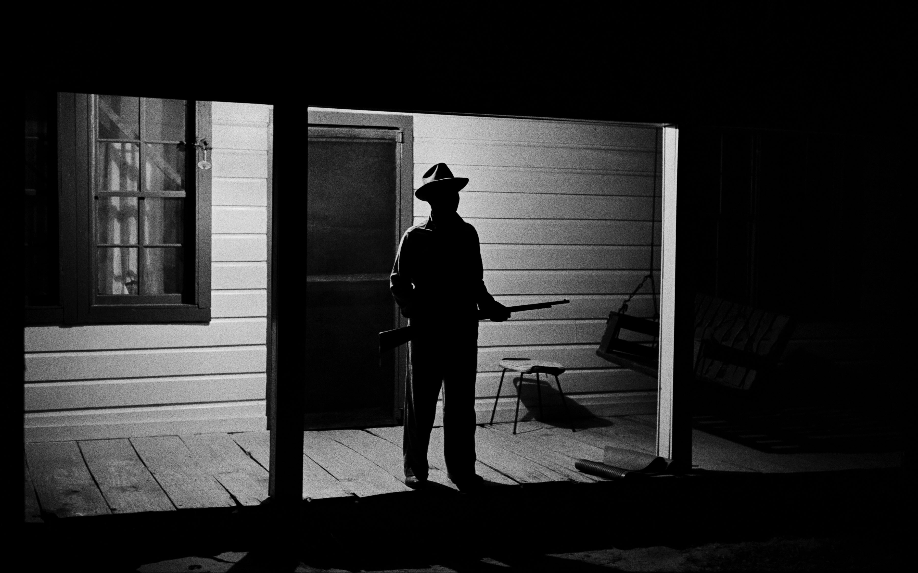 A black-and-white photo of a man wearing a fedora and holding a shotgun standing at the edge of a porch, silhouetted against the porch light behind him.