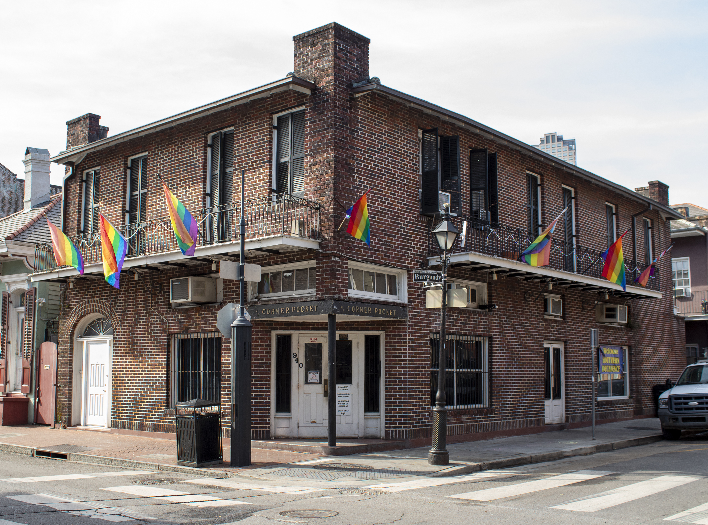 A two-story brick building at a street corner, adorned with multiple rainbow flags. It has white doors and windows, and an outdoor lamp post nearby. The clear sky and urban surroundings create a lively atmosphere.