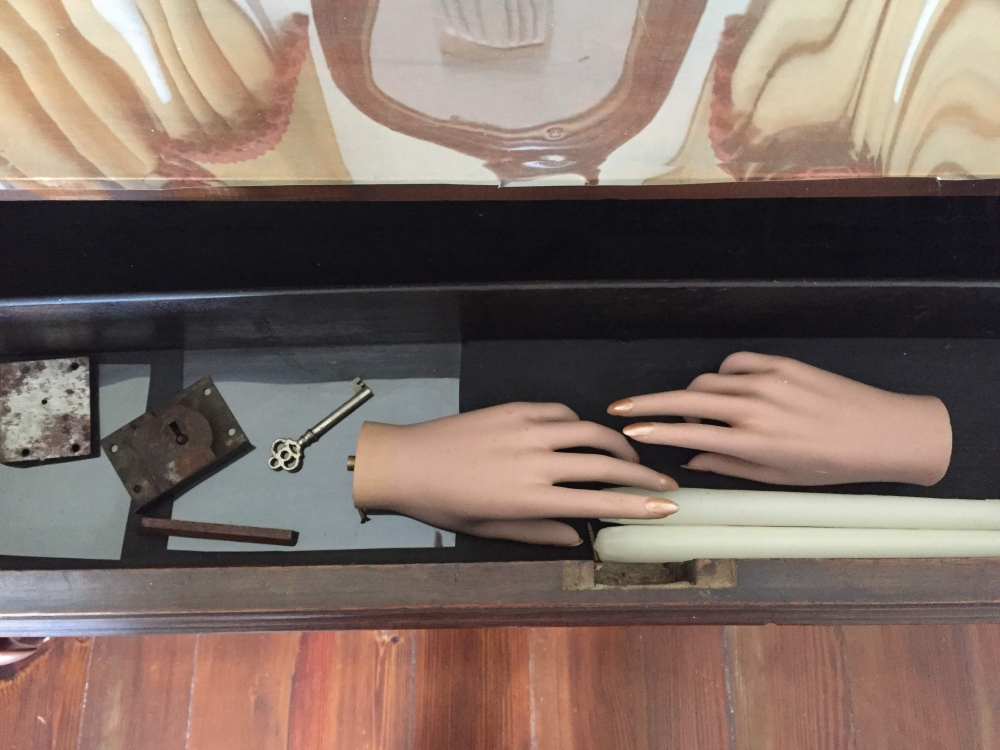 An open drawer containing two mannequin hands, an old key, a lock, a rectangular metal plate, and a white cylindrical object. The drawers wooden interior reflects light, and the floor beneath is wooden.