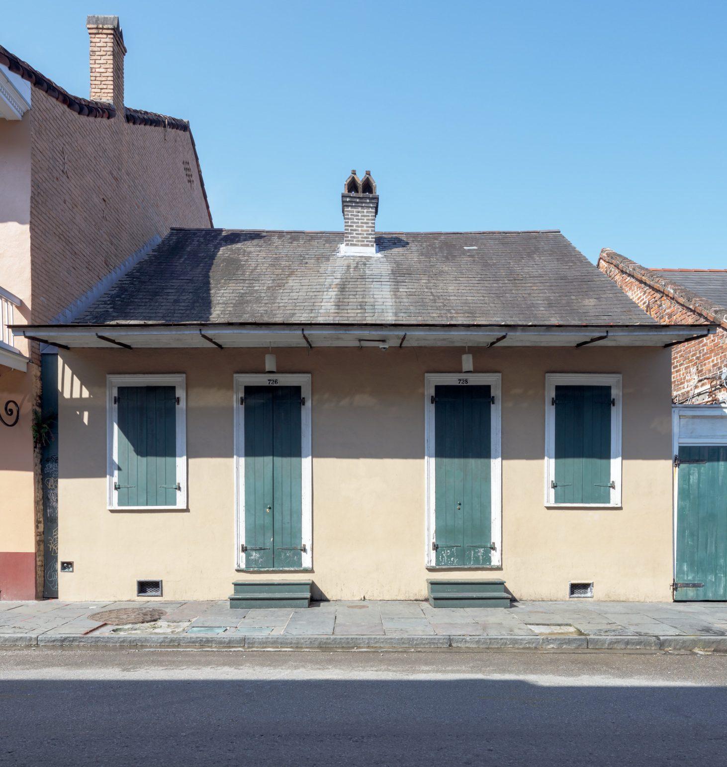 A small, one-story building with three green doors and shutters, located on a quiet street. The building has a simple, sloped roof and is flanked by brick structures. The scene is lit by bright sunlight under a clear blue sky.