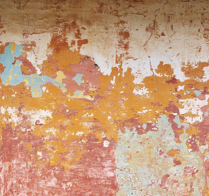 A close-up of a weathered wall with peeling paint in shades of orange, pink, blue, and cream, creating an abstract and textured pattern.