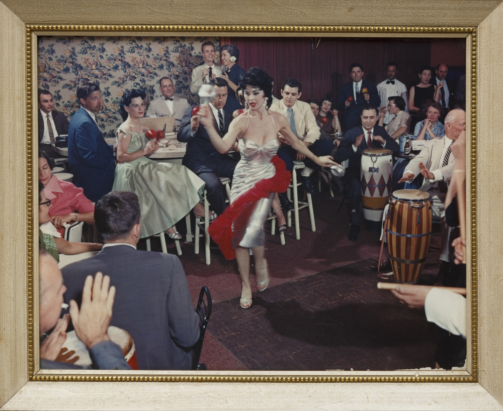 A vibrant scene in a lively club with a dancer in a silver dress performing energetically. Musicians play drums and maracas while an audience watches, some clapping. The atmosphere is festive and joyous, framed in a wooden border.