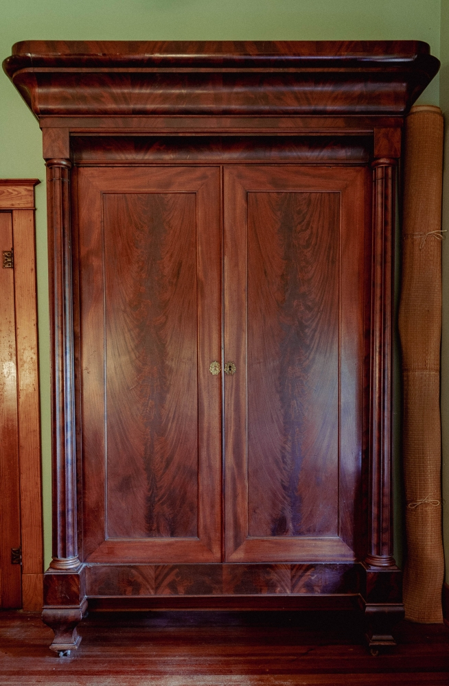 A large, ornate wooden wardrobe with polished finish, featuring double doors and decorative columns. It stands against a green wall, highlighting its rich grain and classical design.
