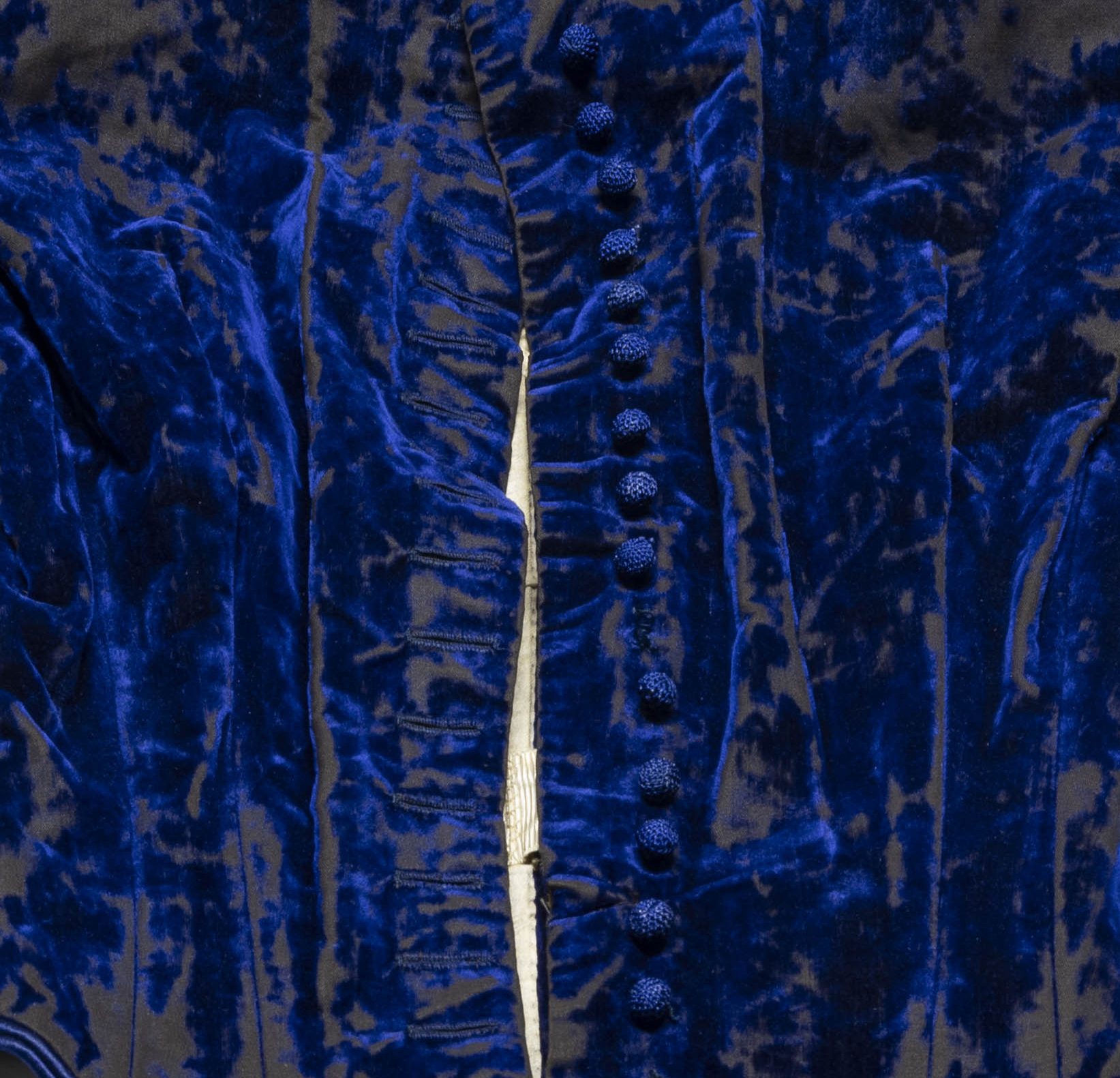 Close up view of a blue velvet bodice, designed by Madame Grignon. A row of buttons can be seen to the right of a center part in the fabric.