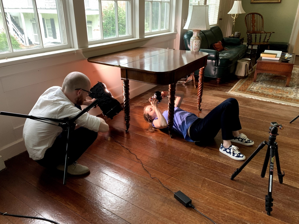 A woman lies on the floor under a wooden table, aiming a camera upwards. A man crouches nearby, assisting with lighting equipment. They are in a brightly lit room with large windows and wooden floors. Various cameras and tripods are around them.