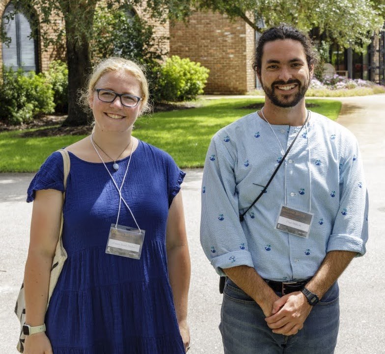 Two people standing outside on a sunny day. They are both smiling and wearing name tags. The woman on the left is in a blue dress, and the man on the right is in a light blue shirt. There are trees and a brick building in the background.