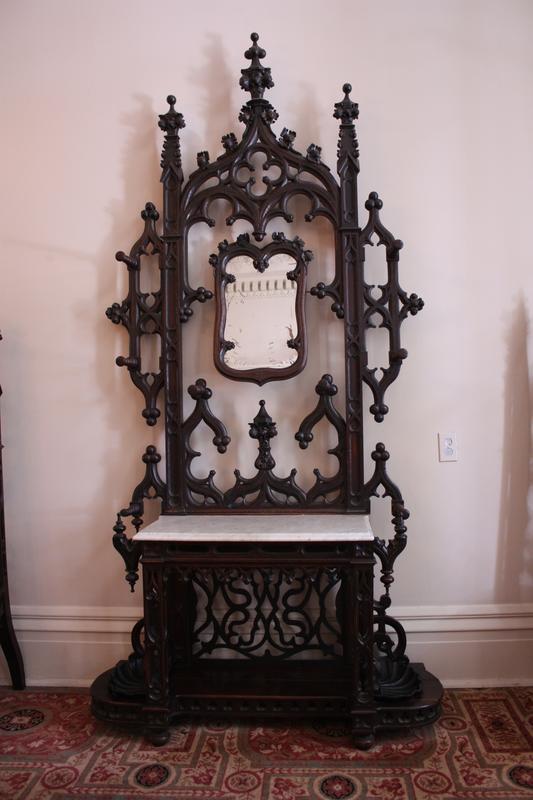 An ornate, dark wood hall tree with intricate carvings, featuring a small mirror at the top and a marble-topped bench below. The furniture stands on a patterned carpet against a white wall.