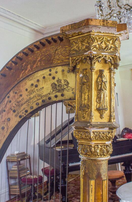 Close-up of a vintage harp with ornate golden carvings, featuring intricate designs of figures and patterns. The soundboard displays a logo and inscriptions. In the background, a grand piano and a stack of music sheets are visible in a well-lit room.