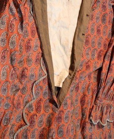 Close-up of a vintage garment with a red paisley pattern on fabric. The clothing includes detailed scalloped edges and gathered fabric on the sleeve, revealing a brown interior lining and an off-white inner material.