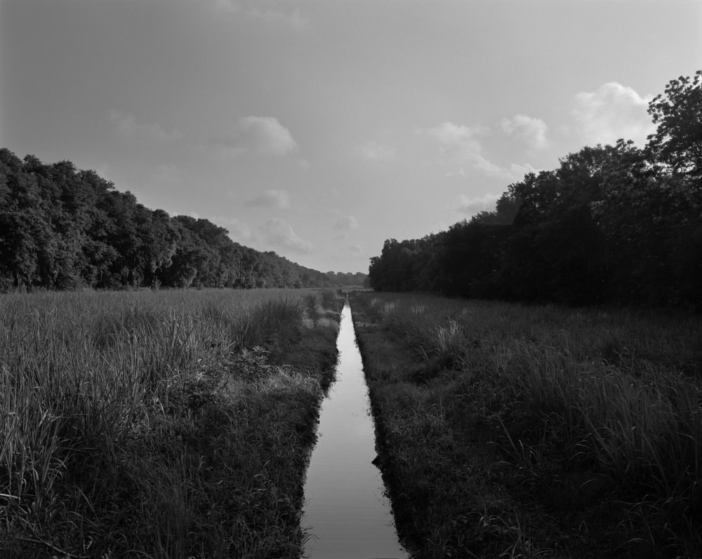 A black and white photo of a narrow canal stretching through a grassy field with trees on either side. The sky is partly cloudy, creating a serene and symmetrical landscape scene.