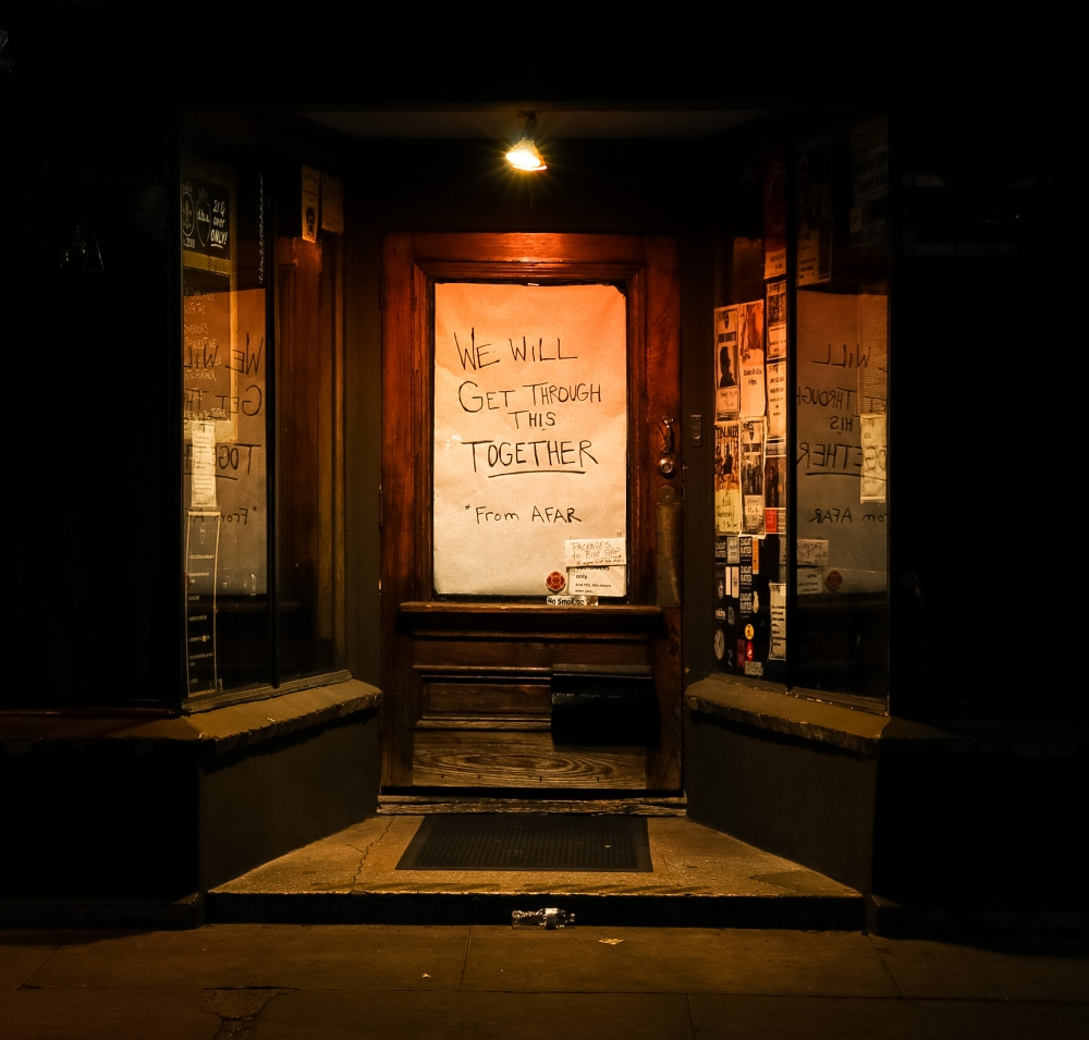 A warmly lit storefront entrance displays a sign on the door that reads, We will get through this together, from afar. Posters line the windows on either side, and the scene is illuminated by a single overhead light.