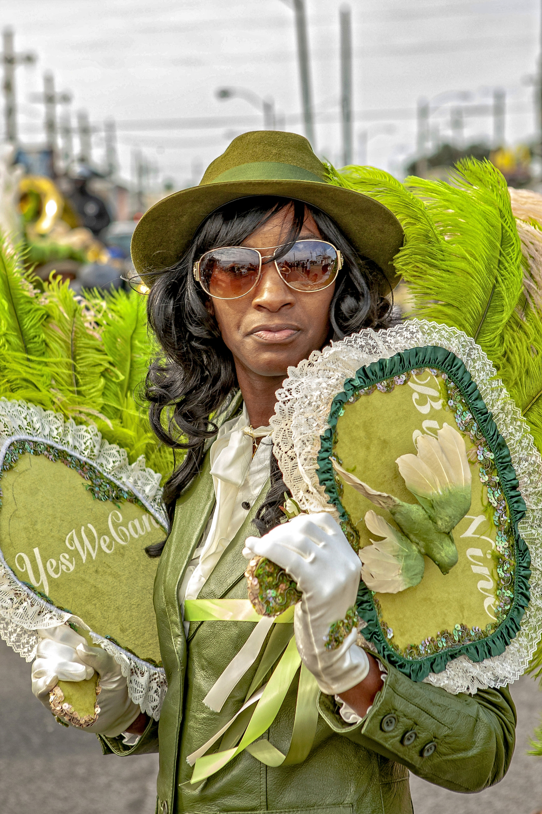 A woman in a stylish green outfit, including a matching hat and aviator sunglasses, holds two ornate fans. One reads "Yes We Can," while the other features a 3D bird decoration. She stands confidently, framed by vibrant green feathers.