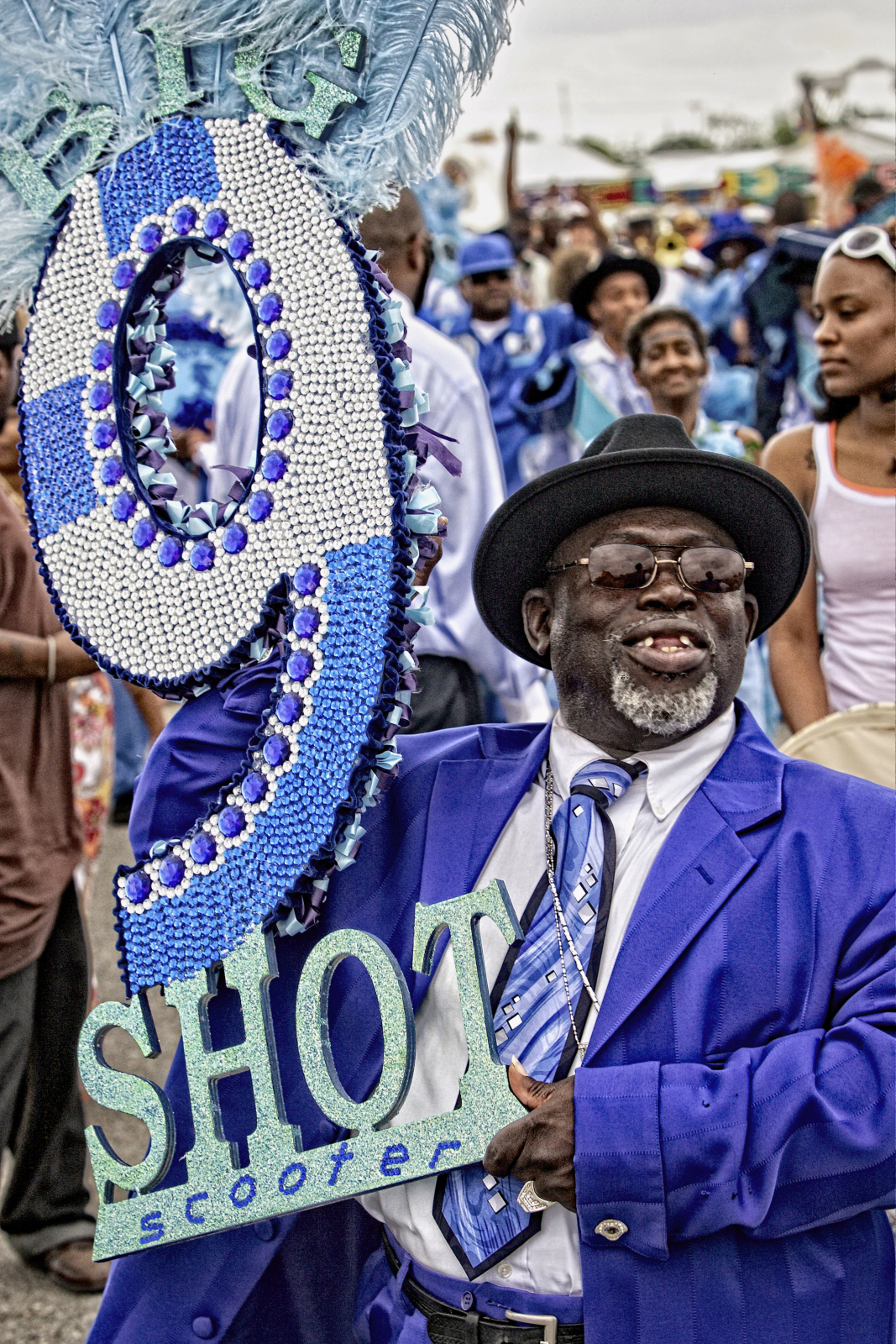A man in a vibrant blue suit, matching tie, and black hat smiles while holding a large, bedazzled number "9" with feathers and the words "SHOT scooter" in glittery letters. A festive crowd in blue attire fills the background.