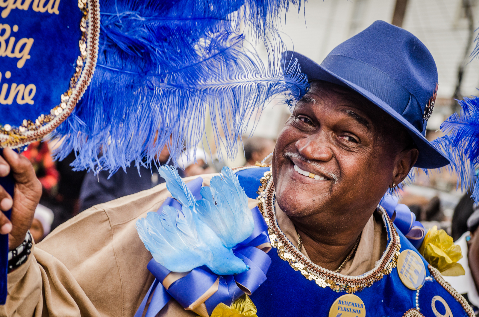 Smiling brightly, a man in a blue hat and decorated vest carries a feathered fan with gold accents. His outfit features intricate details, including a blue bird ornament and tribute buttons.