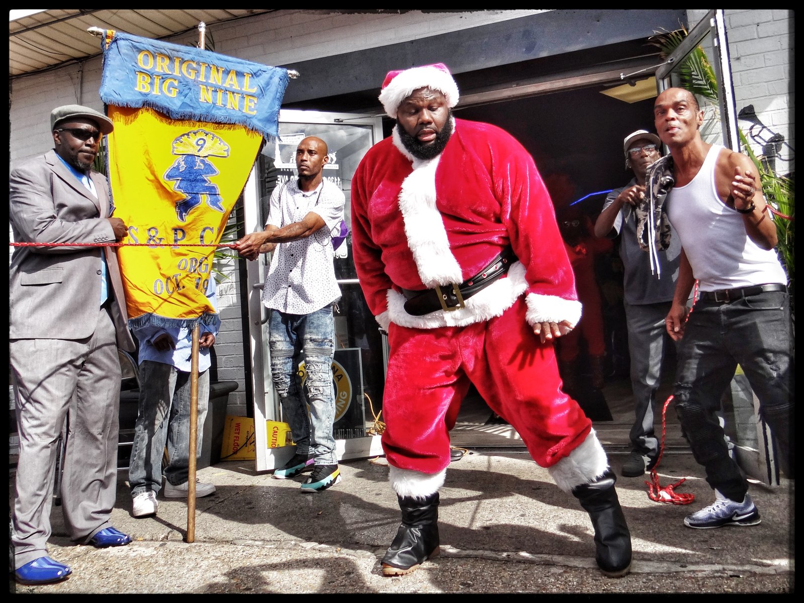A man dressed as Santa dances energetically outside a building, surrounded by casually dressed onlookers and a banner reading "Original Big Nine."