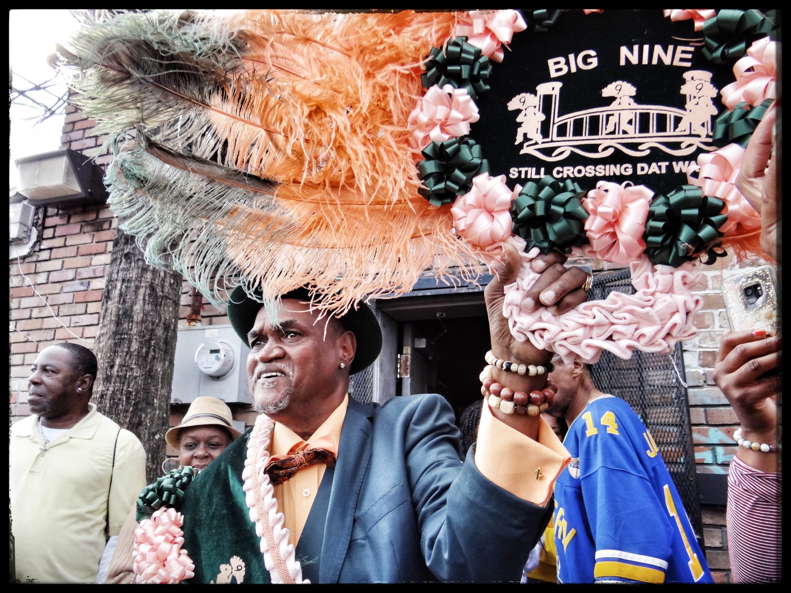 A man in a stylish suit and hat holds up an ornate, feathered parade accessory with “Big Nine” and “Still Crossing Dat Water” written on it.