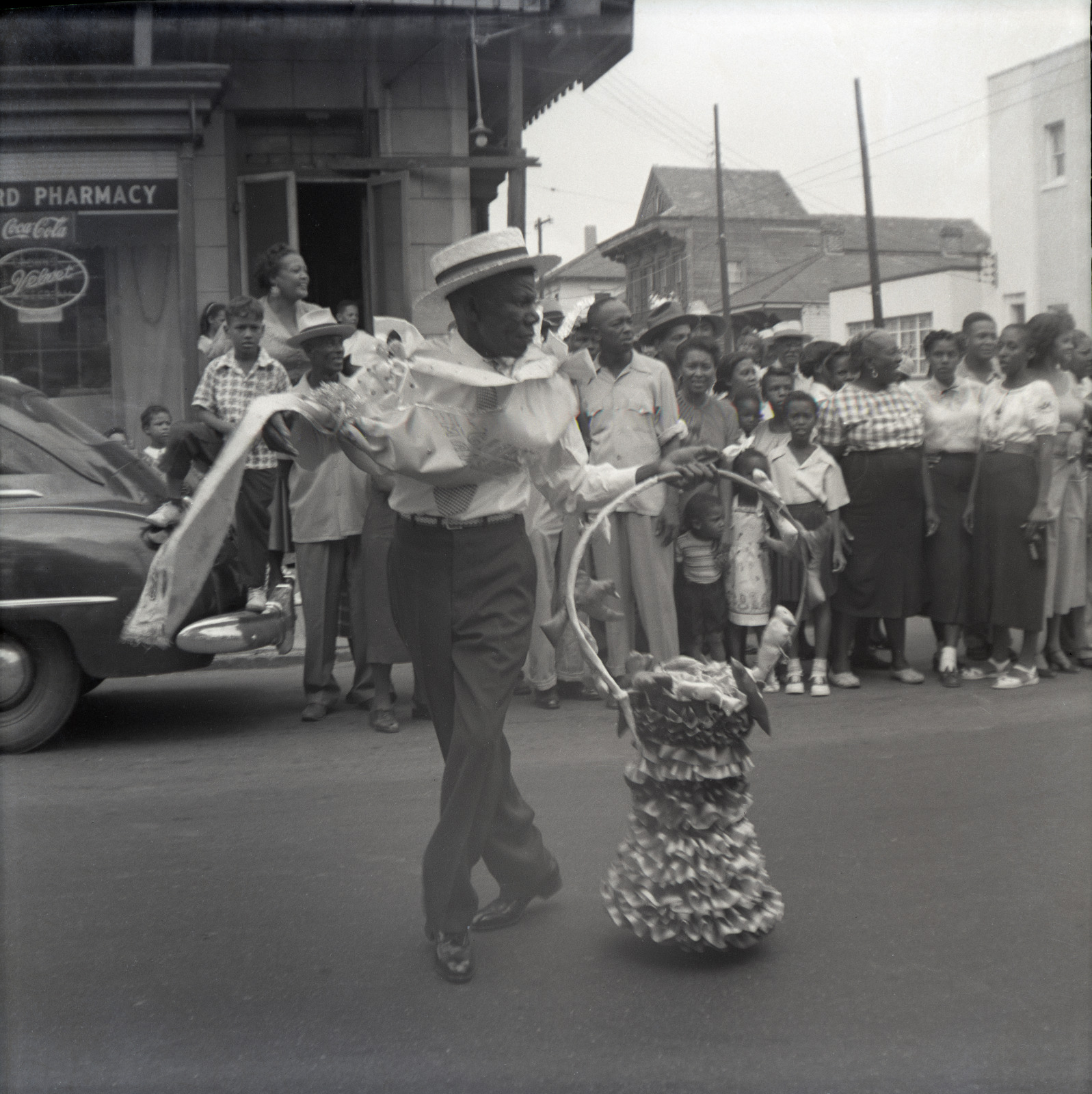 A black and white photo shows a participant in the Original Square Deal Boys parade, who is wearing a white shirt and dark slacks with a straw hat, and is standing with a decorated parade prop.