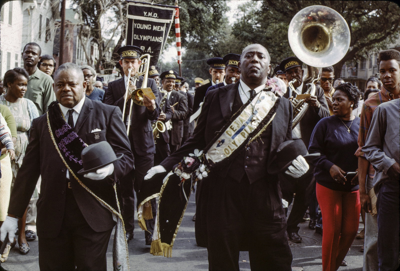 A vintage photo shows members of the YMO Jr. social aid and pleasure club at a funeral second line parade. They are wearing black suits and ties with gold sashes, and are carrying black top hats. A brass band follows behind.