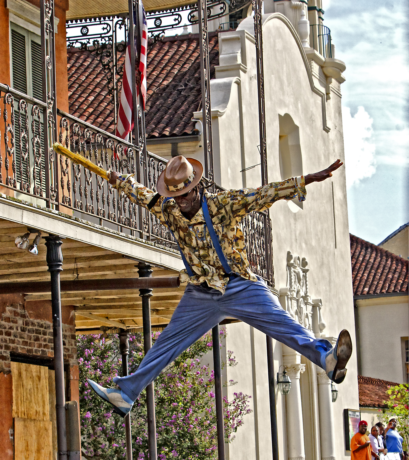 Harold Wilson, wearing a patterned shirt, hat, and blue pants, is captured in midair with his arms and legs outstretched during a second line parade in the French Quarter.