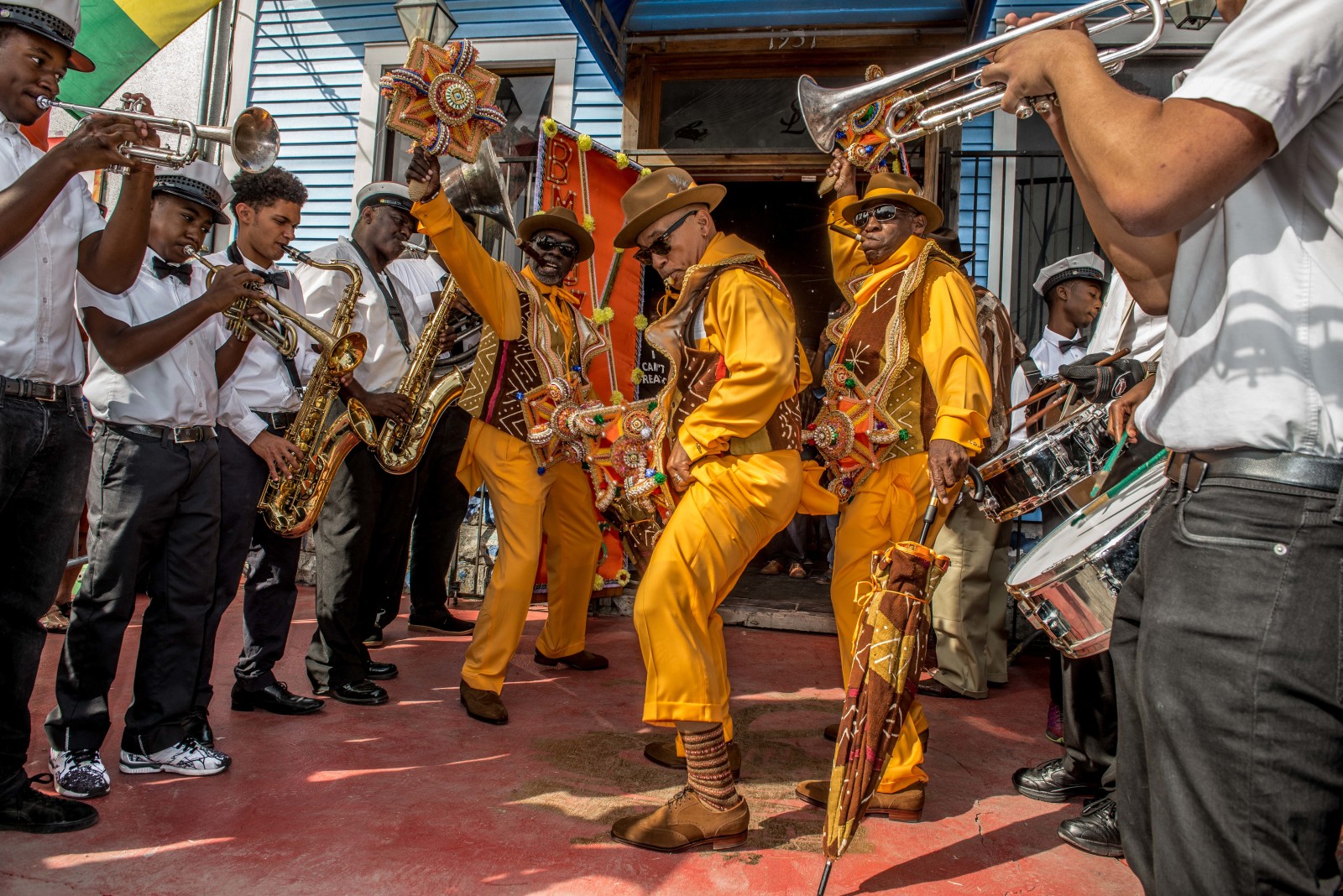 Greg Stafford, Fred Johnson, and Benny Jones Sr. emerge from a doorway in vibrant orange costumes, flanked by brass band musicians on either side.