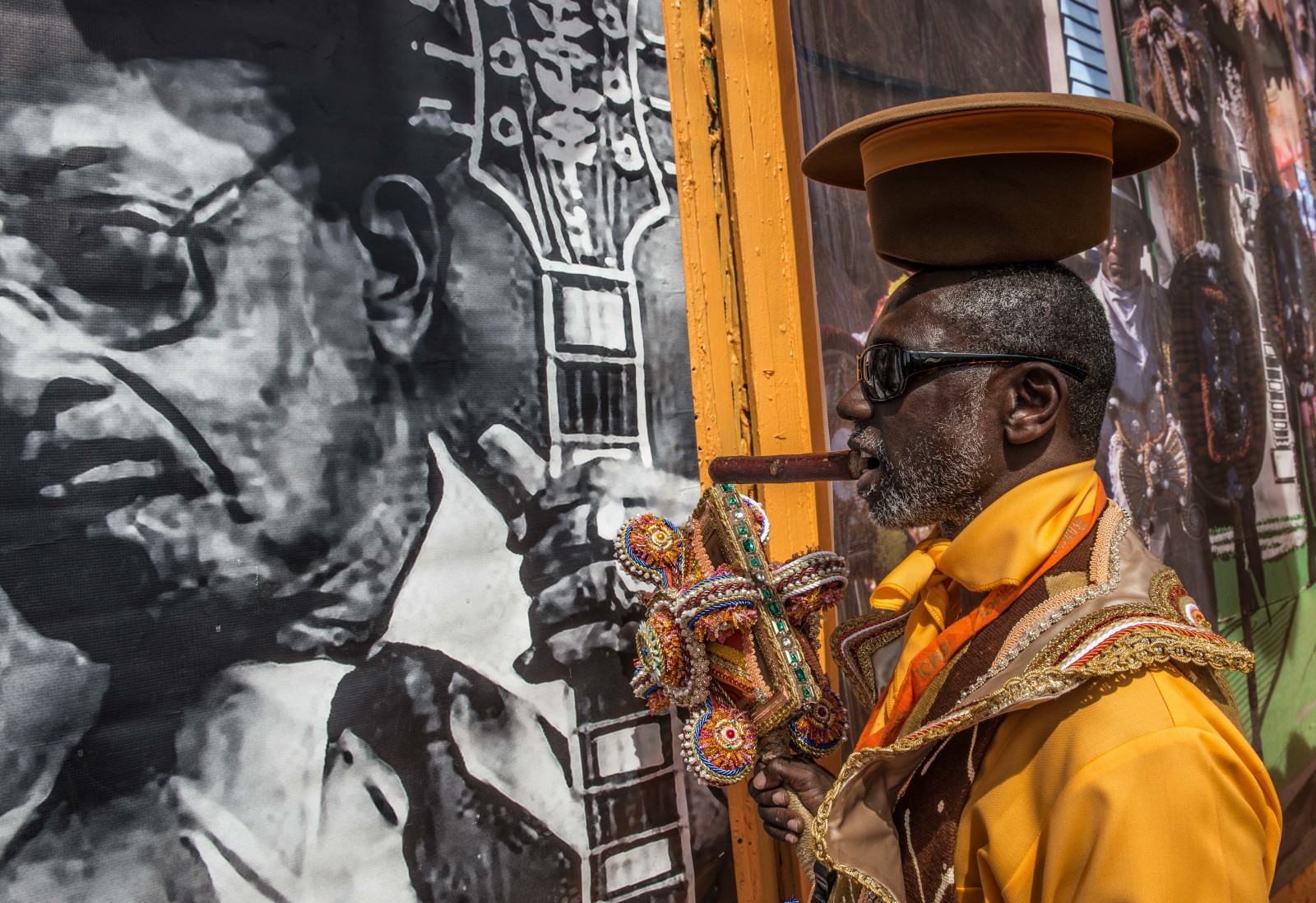 Gregg Stafford, wearing vibrant orange garb and balancing a top hat upside down on his head with a cigar in his mouth, pays tribute to a vintage photo of jazz musician Danny Barker.