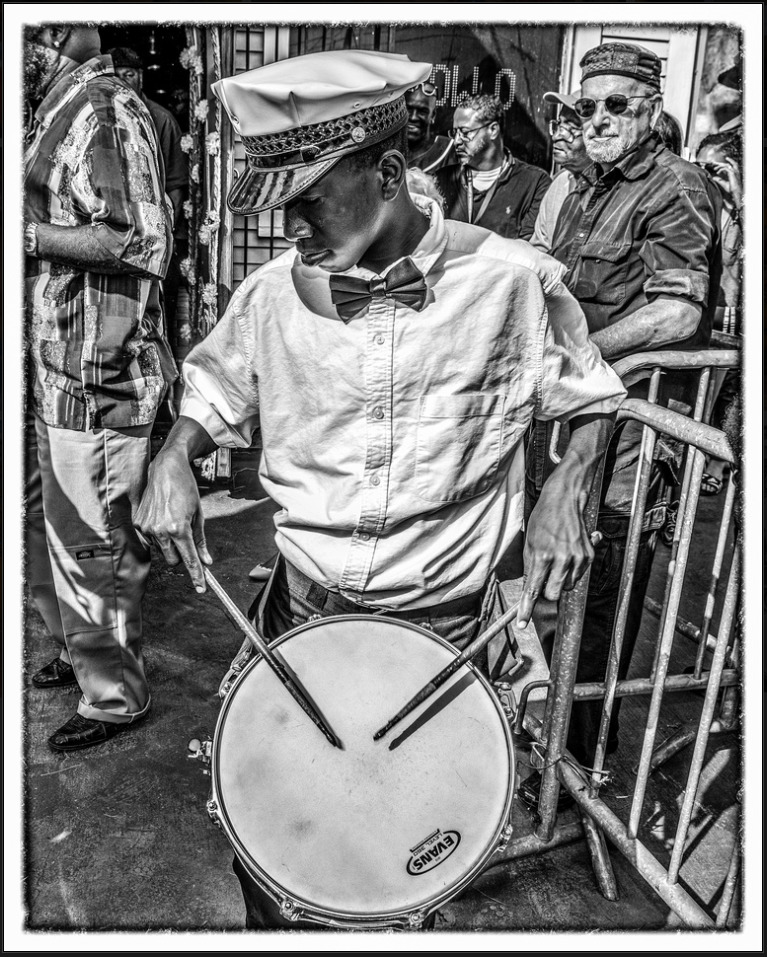 A black and white photo shows Jawansey “NouNou” Ramsey playing the snare drum while wearing a white shirt, white hat, and black bow tie, as part of the New Orleans Young Traditional Brass Band.