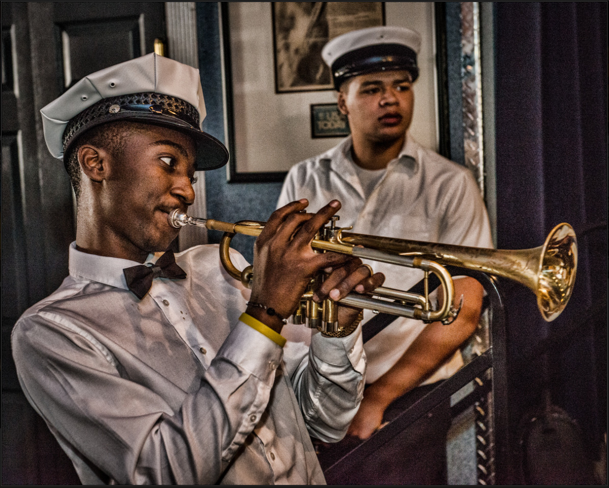 Emmanuel Mitchell Jr. plays trumpet while dressed in a white hat, white dress shirt, and black bow tie as part of the New Orleans Young Traditional Brass Band. Musician Aurélien Barnes stands in the background.