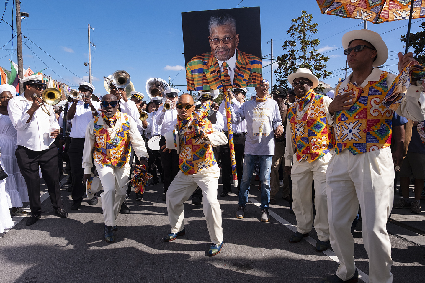 Members of the Black Men of Labor parade down the street, while carrying a memorial portrait of a deceased member. They are wearing vibrant, multicolored patterned vests, white pants, and white hats, with a brass band following behind.