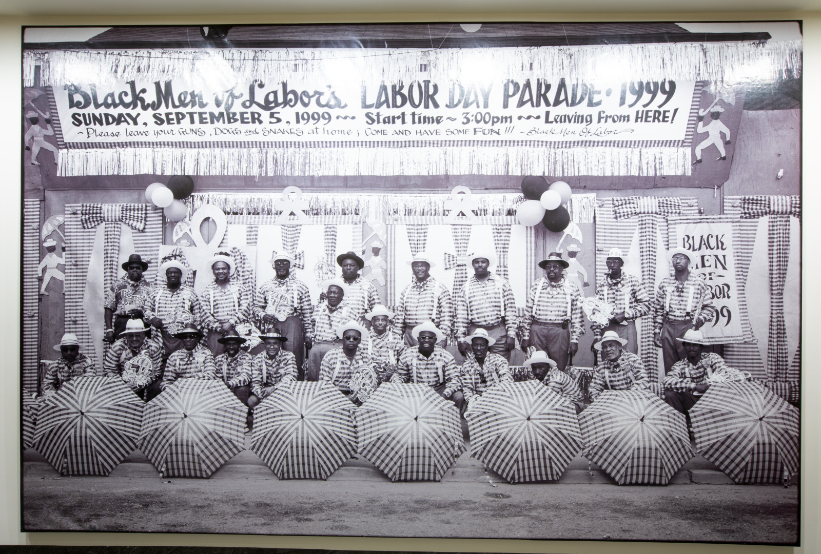 A black and white photo shows members of the Black Men of Labor posing under a large banner that reads "Black Men of Labor Labor Day Parade 1999". Each member is wearing plaid suits with white hats, with matching parasols on the ground at their feet.