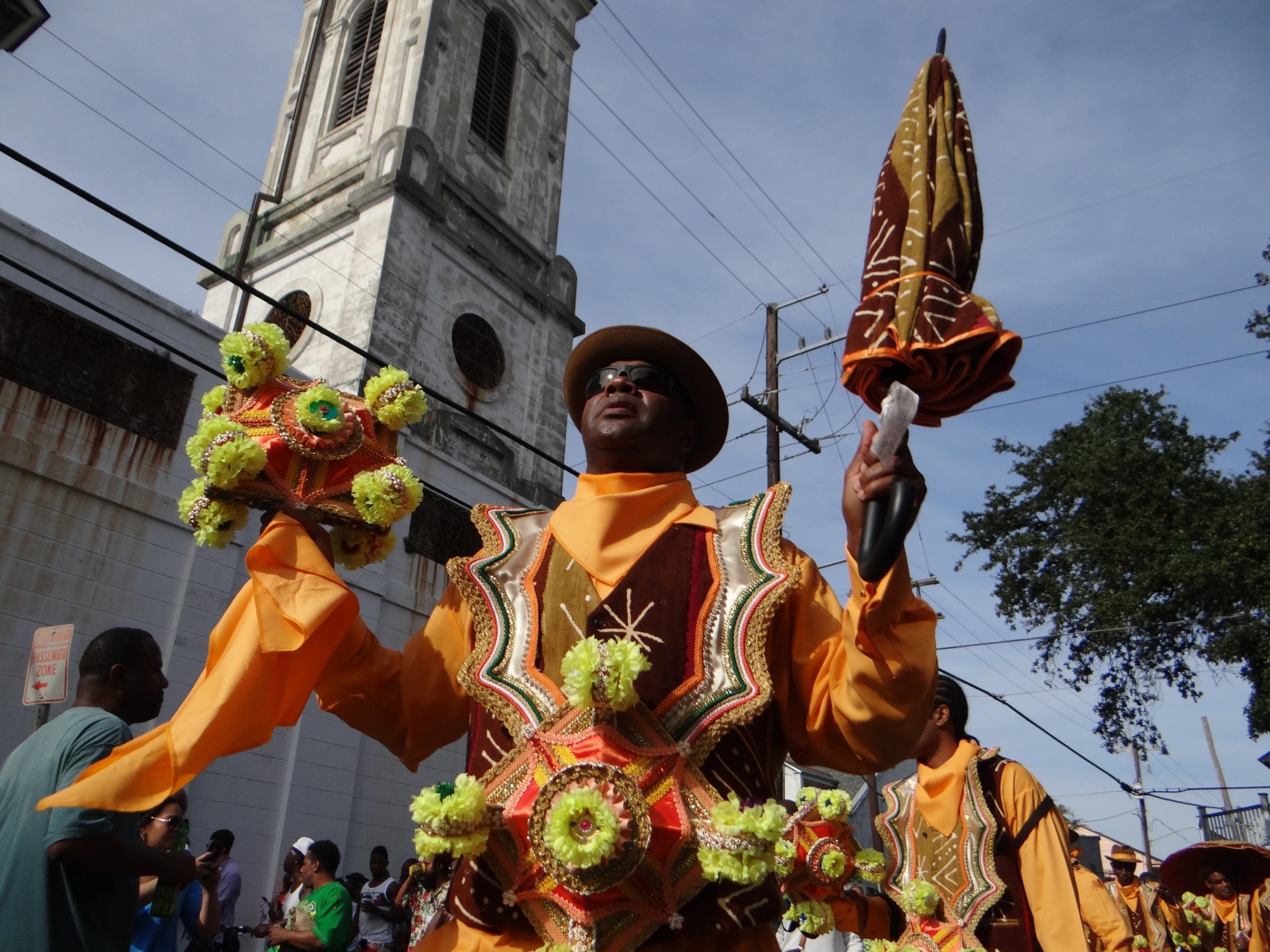 Bruce “Sunpie” Barnes wears an ornate red and orange costume at a second line parade, with a folded parasol in one hand and a flower-decorated prop in the other. A church steeple is visible in the background.