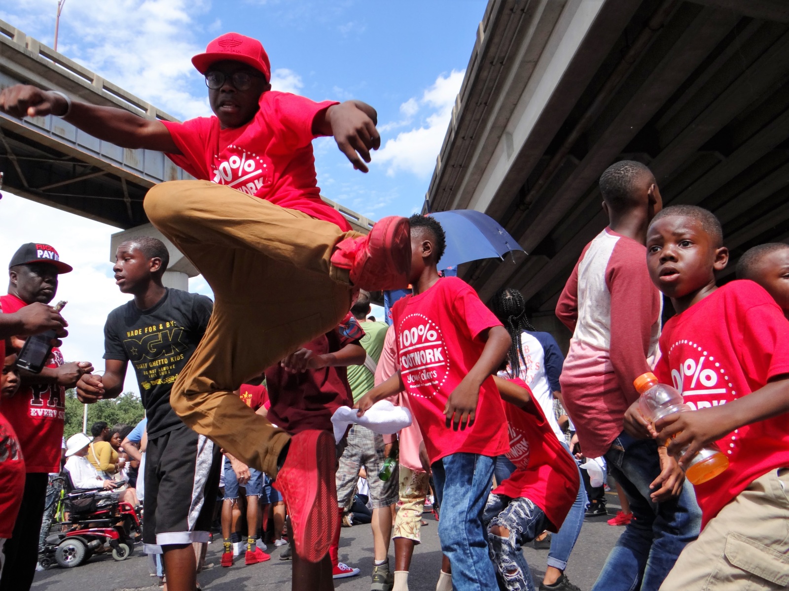 A group of kids at the Family Ties second line parade, wearing red t-shirts, pants, and shoes, dance under the Claiborne overpass. One of them is photographed several feet off the ground.