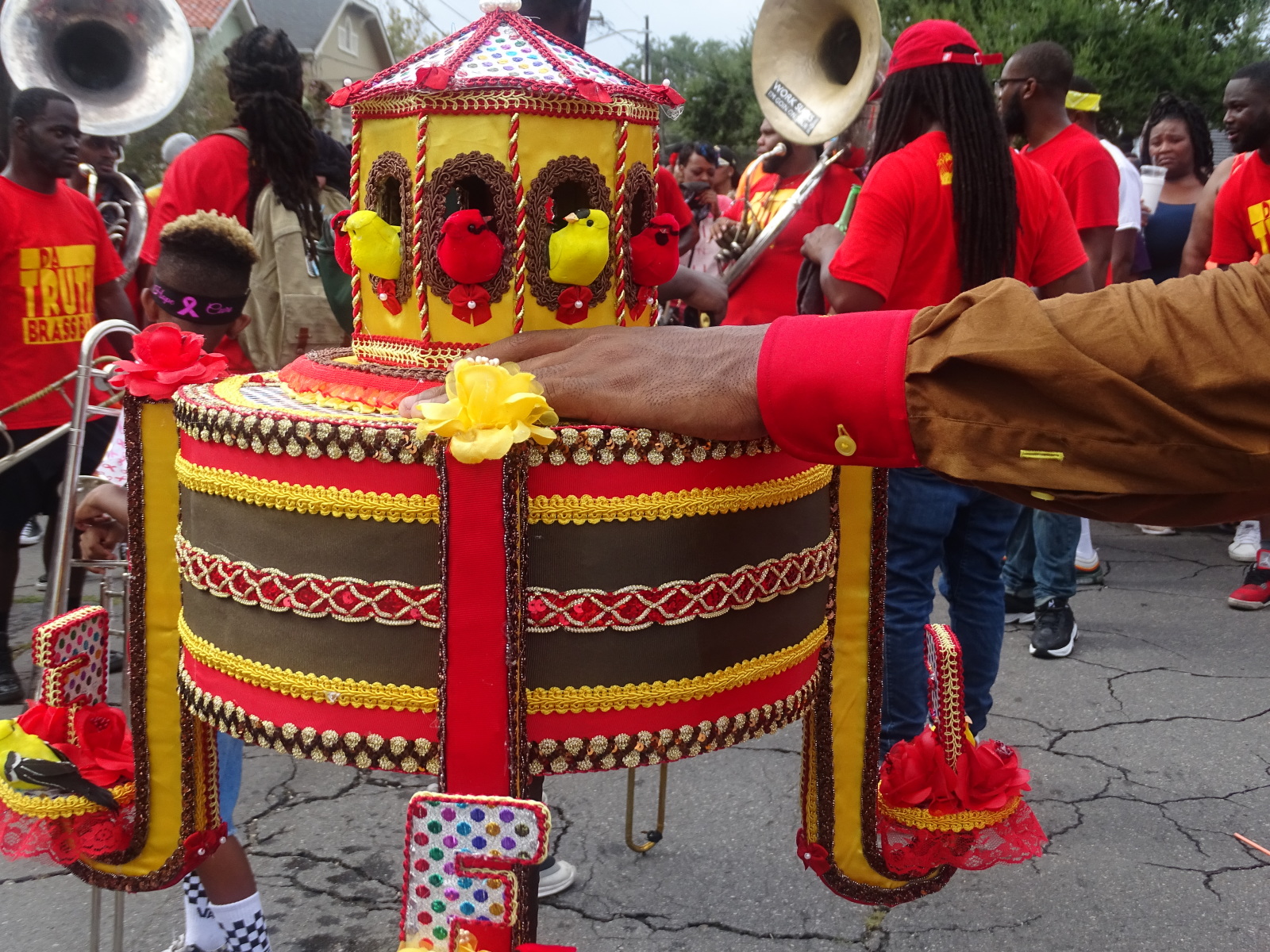 Red and yellow embroidered artwork created by Derrick Santa Cruz for Footwerk Family is on display in front of a brass band at a second line parade.